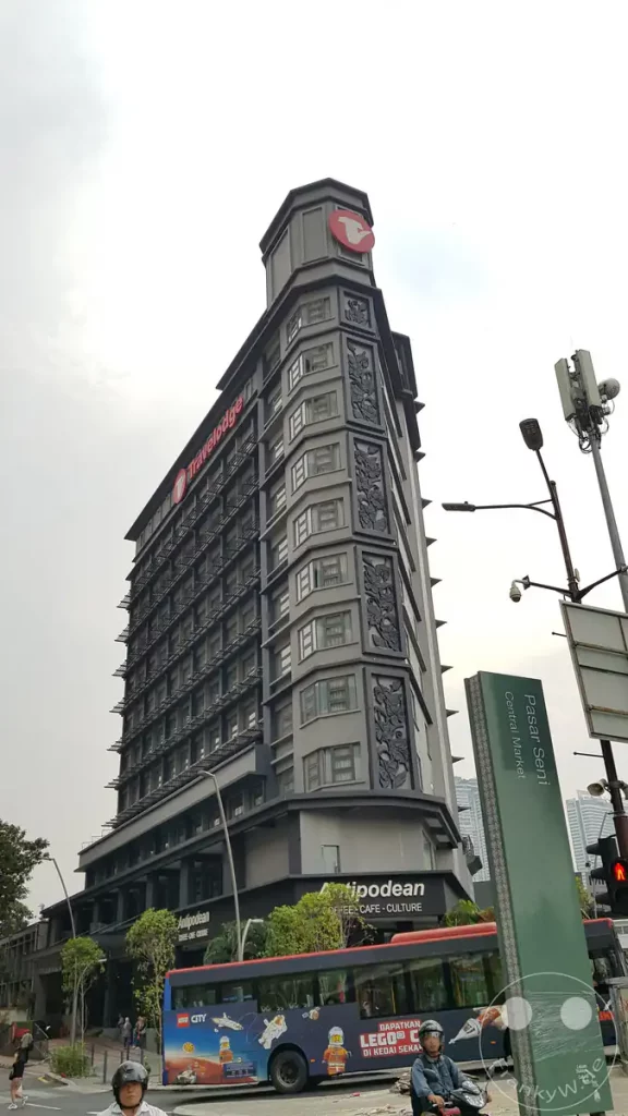 Froschperspektive auf das markante, schmale Gebäude des Travelodge City Centre Hotels mit verzierten grauen Reliefelementen, einem Antipodean Cafe im Erdgeschoss und einem Linienbus an einer Straße nahe Pasar Seni in Kuala Lumpur, Malaysia.