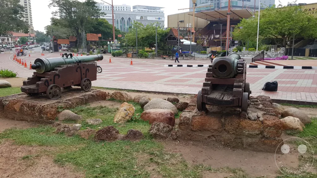 Zwei historische Kanonen auf Holzlafetten vor den Überresten der Festung A Famosa in Malakka, Malaysia, aufgestellt auf steinernen Sockeln mit Blick auf einen gepflasterten Platz und das Einkaufszentrum Dataran Pahlawan im Hintergrund.