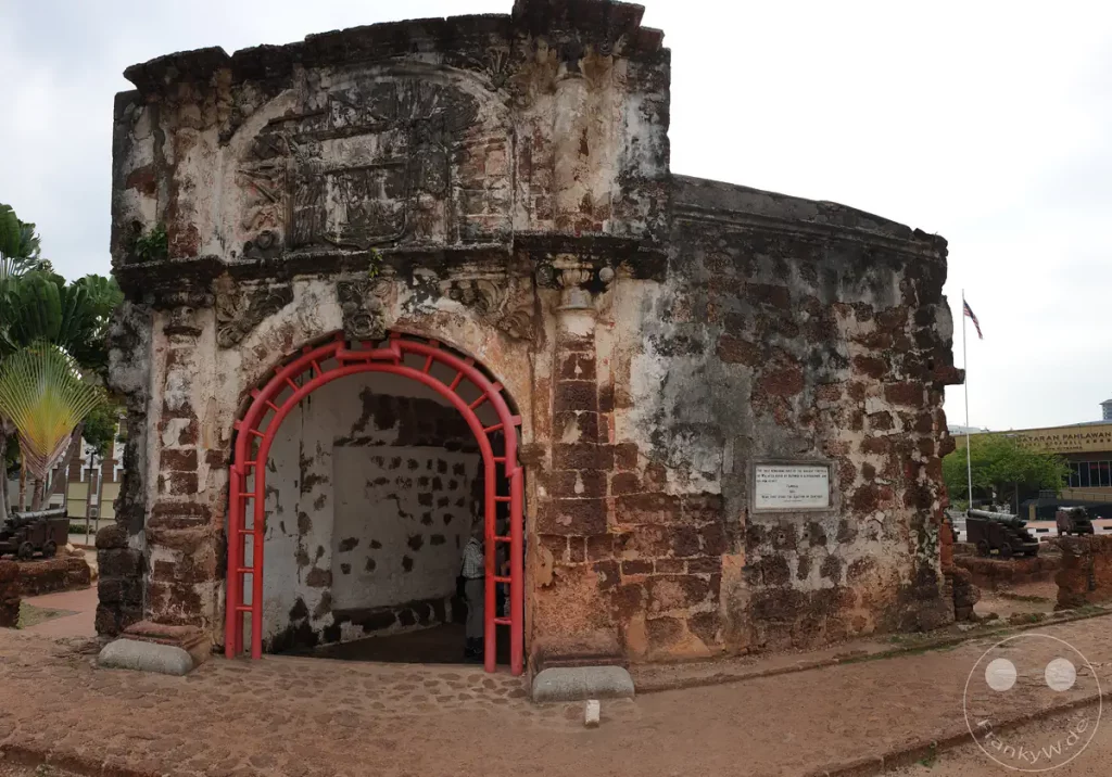 Blick auf das historische Festungstor Porta de Santiago der ehemaligen portugiesischen Festung A Famosa in Malakka, Malaysia, mit seinen verwitterten Laterit-Steinmauern, einem rötlichen Torbogen und alten Kanonen im Hintergrund.