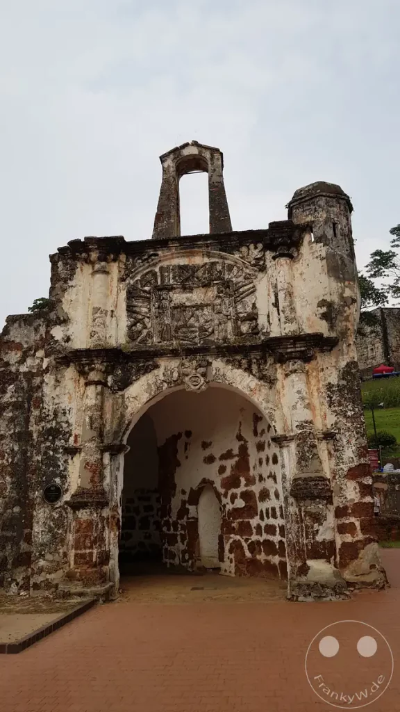 Frontalansicht des historischen, verwitterten Steintors Porta de Santiago der ehemaligen portugiesischen Festung A Famosa in Malakka, Malaysia, mit einem markanten Torbogen, Reliefs über dem Eingang und einer offenen Glockenöffnung an der Spitze.
