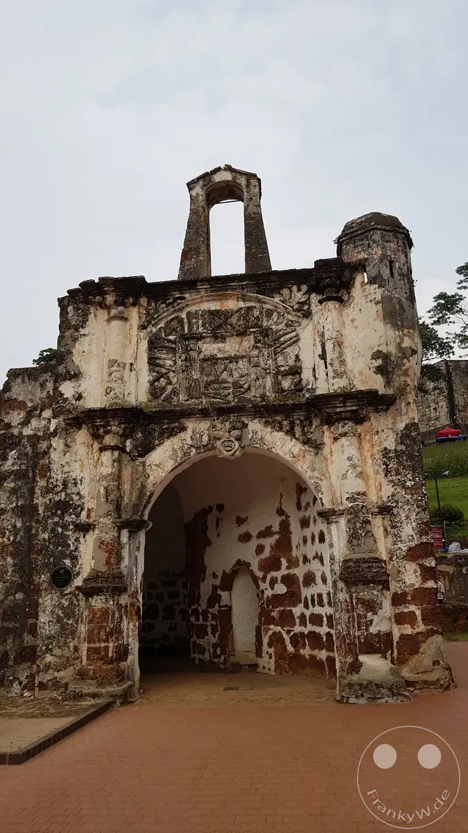 Frontalansicht des historischen, verwitterten Steintors Porta de Santiago der ehemaligen portugiesischen Festung A Famosa in Malakka, Malaysia, mit einem markanten Torbogen, Reliefs über dem Eingang und einer offenen Glockenöffnung an der Spitze.