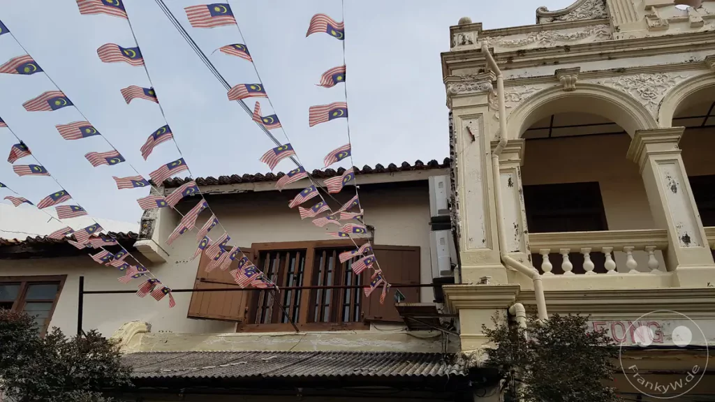Zahlreiche kleine malaysische Flaggen hängen an Girlanden gespannt über einer historischen Straße in Malakka vor der Kulisse traditioneller Gebäude mit kunstvollen Fassaden.