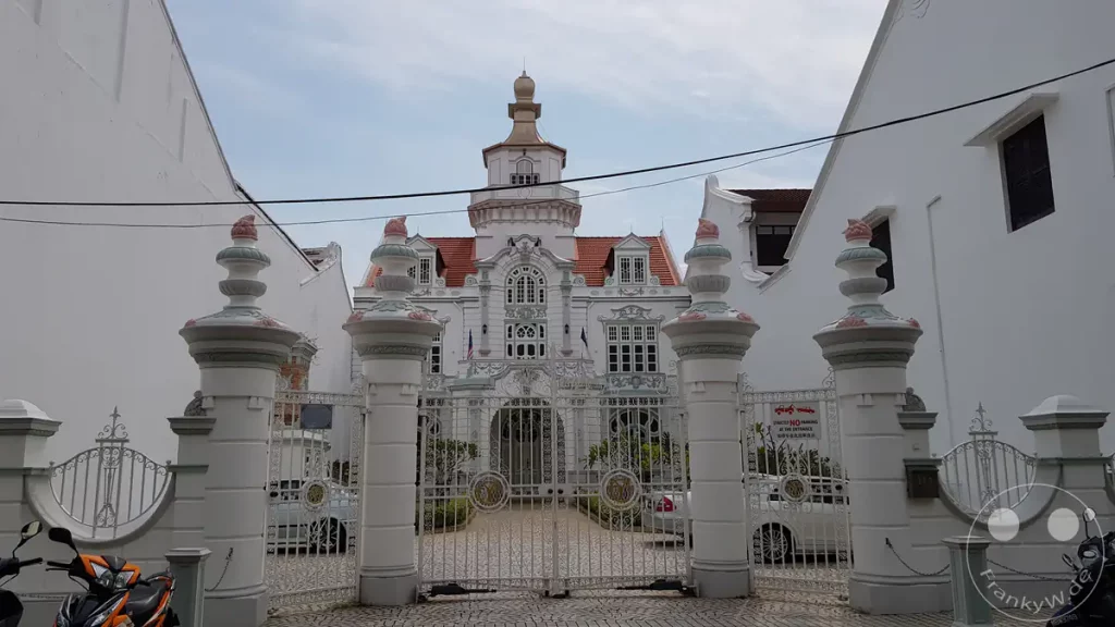 Das historische Chee-Herrenhaus (Chee Mansion) in Malakka mit seiner prachtvollen weißen Fassade, verzierten Toren und einem auffälligen Turm im barocken Kolonialstil unter bewölktem Himmel.
