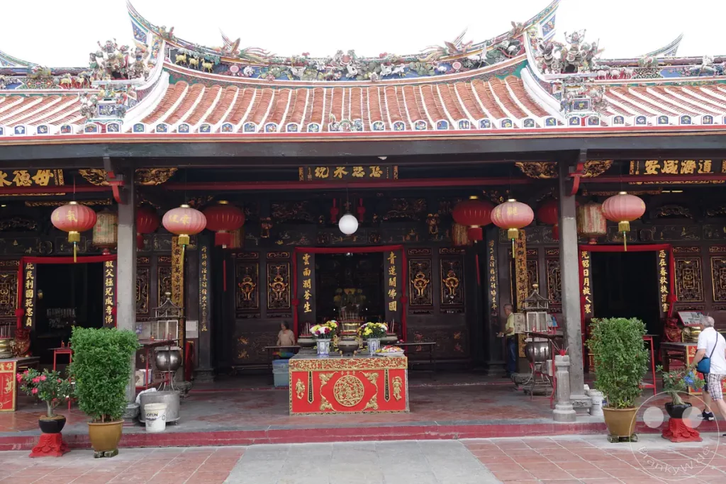 Der reich verzierte Eingangsbereich des historischen Cheng Hoon Teng Tempels in Malakka, Malaysia, mit traditionellen roten Lampions, vergoldeten Schriftzeichen auf schwarzen Holzsäulen, einem Opfertisch im Zentrum und kunstvollen Skulpturen auf dem geschwungenen Ziegeldach.
