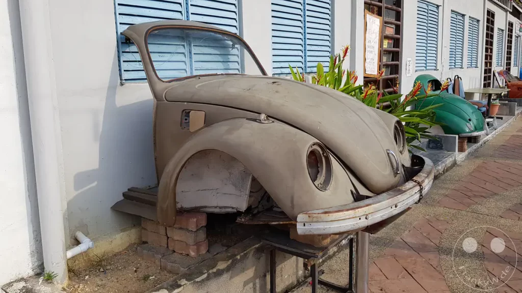 Nahaufnahme einer dekorativen, grauen Oldtimer-Front eines VW Käfers aufgebockt vor einem Gebäude mit hellblauen Fensterläden an der Uferpromenade des Malakka-Flusses am Discovery Cafe in Malakka, Malaysia.