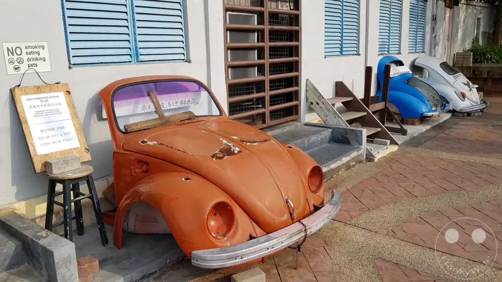 Blick auf mehrere dekorative, bunte Oldtimer-Fronten von VW Käfern in Orange, Blau und Weiß vor hellblauen Fensterläden an der Uferpromenade des Malakka-Flusses am Discovery Cafe in Malakka, Malaysia.