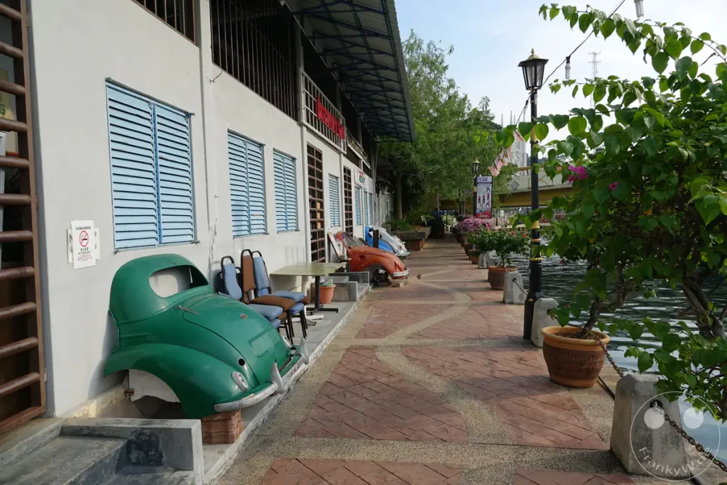 Blick entlang der gepflasterten Uferpromenade des Malakka-Flusses am Discovery Cafe in Kampung Jawa, Malakka, Malaysia, mit dekorativen, bepflanzten Oldtimer-Fronten von VW Käfern vor hellblauen Fensterläden.