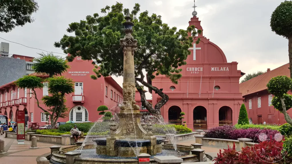 Blick auf den historischen Dutch Square in Malakka, Malaysia, mit dem plätschernden Queen Victoria Brunnen im Vordergrund, der markanten roten Christ Church und der Melaka Art Gallery unter einem bewölkten Himmel.