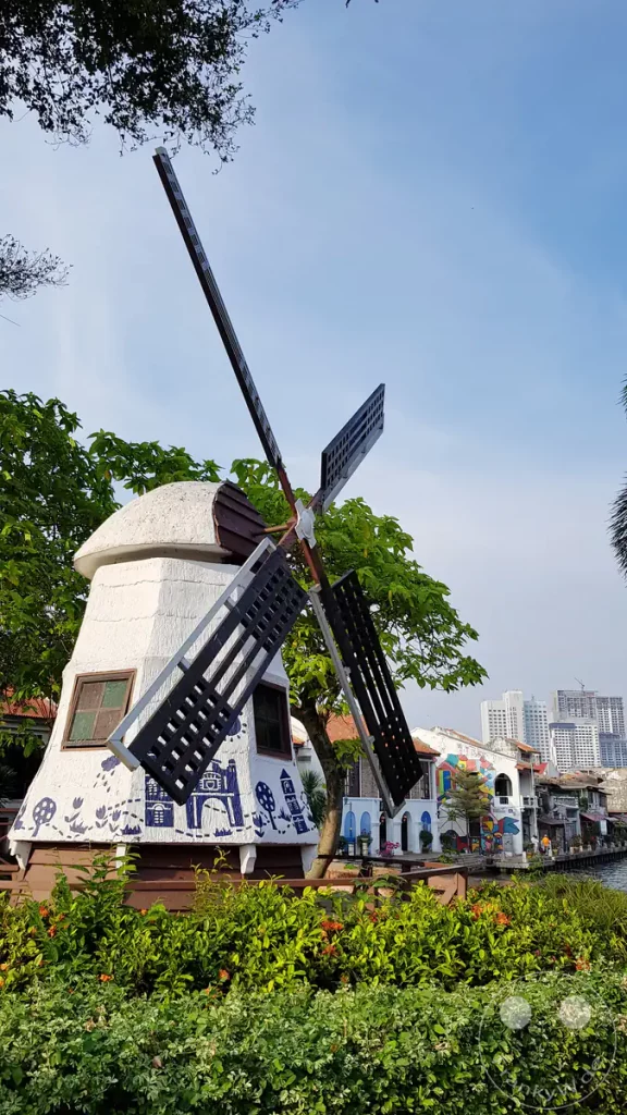 Blick auf die nachgebaute, weiß-blaue holländische Windmühle am Dutch Square in Malakka, Malaysia, umgeben von grünen Hecken mit Blick auf historische Gebäude an der Flussuferpromenade und moderne Hochhäuser in der Ferne.