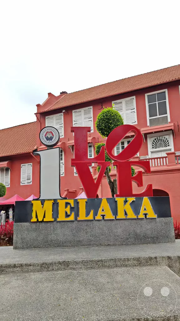 Nahaufnahme des rot-gelben Schriftzugs „I Love Melaka“ auf einem Steinsockel am historischen Dutch Square in Malakka, Malaysia, mit dem markanten roten Stadthuys-Gebäude im Hintergrund.