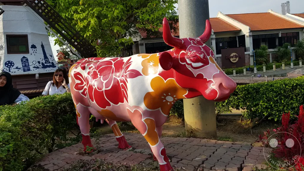 Nahaufnahme einer rot-weiß gemusterten Kuh-Skulptur mit Blumenmotiven auf einem Pflasterweg am Dutch Square in Malakka, Malaysia, mit einer nachgebauten Windmühle links und dem Hard Rock Cafe im Hintergrund.