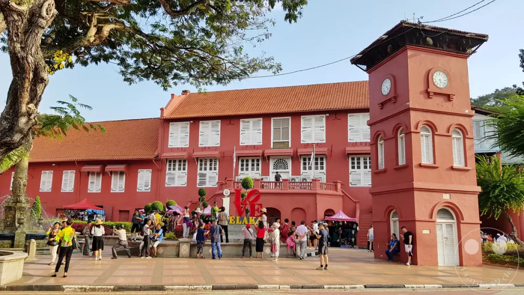 Blick auf den historischen Dutch Square in Malakka, Malaysia, mit dem markanten roten Uhrturm, dem Stadthuys-Gebäude mit weißen Fensterläden, einem „I Love Melaka“-Schriftzug und zahlreichen Touristen auf dem Platz.