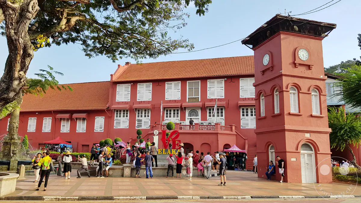 Blick auf den historischen Dutch Square in Malakka, Malaysia, mit dem markanten roten Uhrturm, dem Stadthuys-Gebäude mit weißen Fensterläden, einem „I Love Melaka“-Schriftzug und zahlreichen Touristen auf dem Platz.