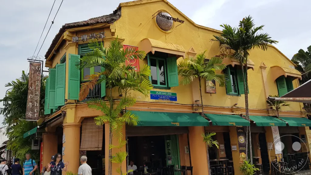 Das markante gelbe Geographer Cafe in Malakka an einer Straßenecke mit grünen Fensterläden, Markisen und Palmen vor der historischen Shophouse-Fassade im Kolonialstil.