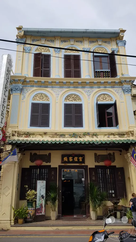 Die historische, mehrstöckige Fassade des Hotel Puri in Malakka im traditionellen Peranakan-Stil mit gelb-blauen Verzierungen, dunklen Holzfensterläden und chinesischen Schriftzeichen am Eingang.
