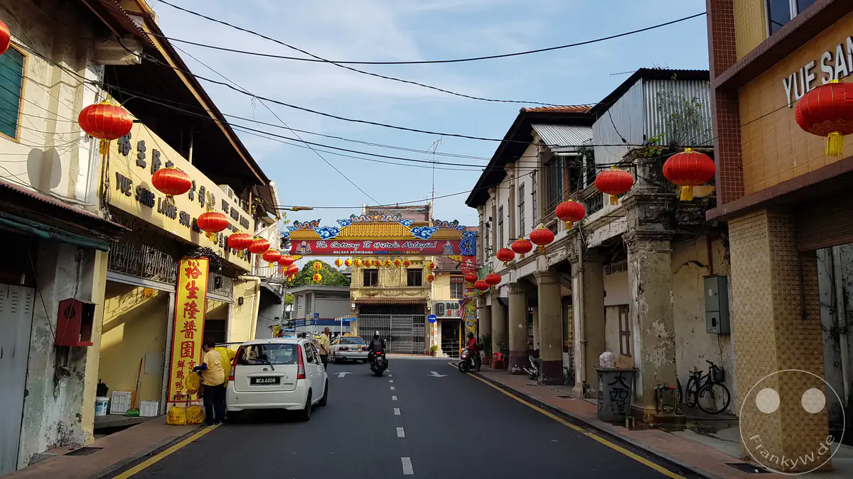 Blick entlang der Jalan Kampung Pantai in Malakka, Malaysia, mit historischen Peranakan-Shophouses im Kolonialstil, roten Lampions und einem dekorativen blauen Torbogen im Hintergrund mit der Aufschrift „The Gateway to Historic Malaysia!“.