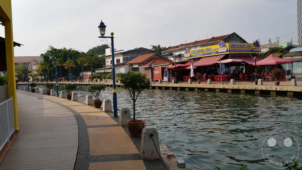 Blick auf die Uferpromenade entlang des Malakka-Flusses in Kampung Jawa, Malakka, Malaysia, mit dem Restaurant „Line Clear Kg. Jawa“, Straßenlaternen und bepflanzten Blumentöpfen am Flussufer.