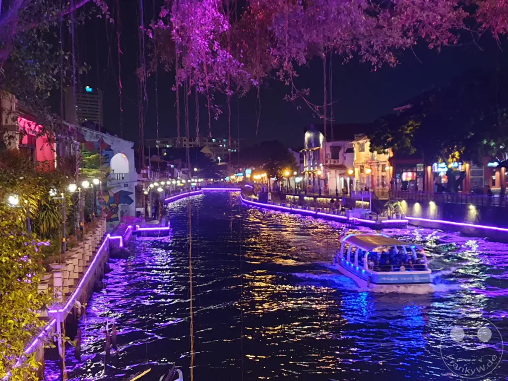 Nachtaufnahme des hell erleuchteten Malakka-Flusses in Malakka, Malaysia, mit einem vorbeifahrenden Boot der Melaka River Cruise, lila illuminierten Uferpromenaden und hängenden Ästen im Vordergrund.