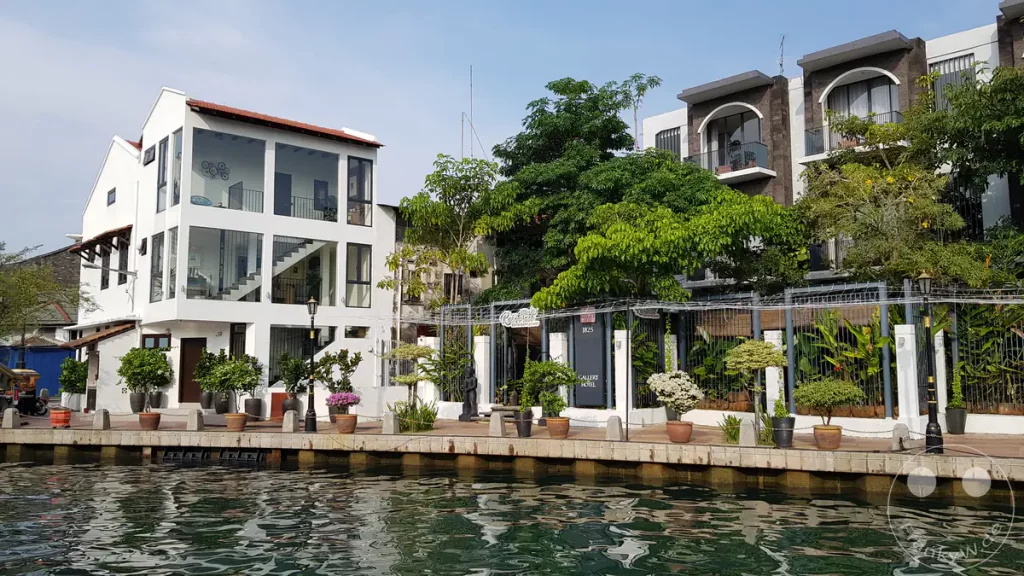 Blick vom Fluss auf das moderne weiße Gebäude des Backyard Rio Melaka und die begrünte Uferpromenade des historischen 1825 Gallery Hotels am Malakka-Fluss in Malakka, Malaysia.