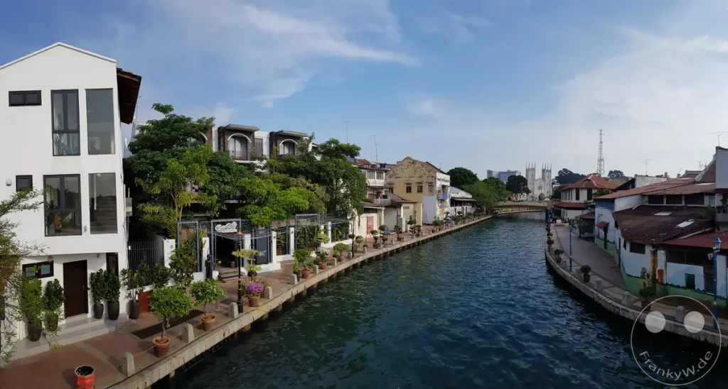Panoramablick auf den geschwungenen Malakka-Fluss in Malakka, Malaysia, mit der gepflasterten Uferpromenade, dem 1825 Gallery Hotel auf der linken Seite und den weißen Türmen der St.-Francis-Xavier-Kirche in der Ferne.