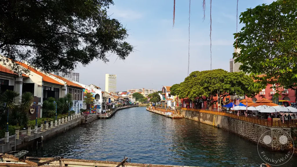 Blick von einer Brücke auf den Malakka-Fluss in Malakka, Malaysia, gesäumt von historischen Shophouses auf der linken Seite, der roten Ufermauer am Roten Platz mit Restaurantzelten auf der rechten Seite und Bäumen unter blauem Himmel.