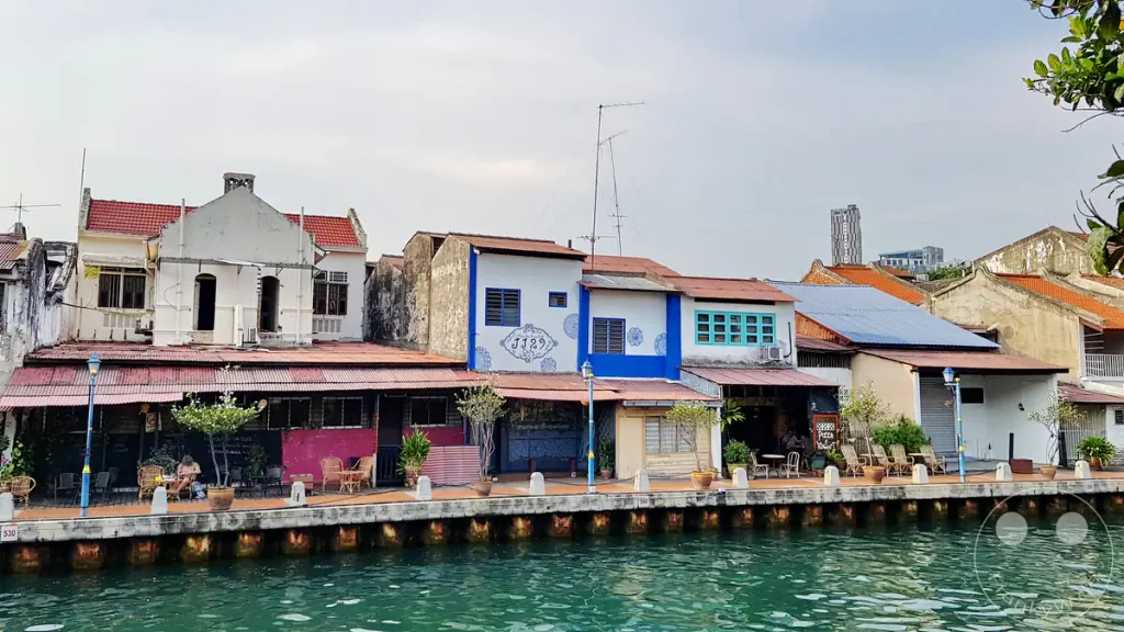 Blick über den Malakka-Fluss auf historische Shophouses an der Uferpromenade in Malakka, Malaysia, darunter das blau-weiße Gebäude mit der Aufschrift „JJ29“, kleine Cafés mit Sitzgelegenheiten im Freien und traditionelle Ziegeldächer.