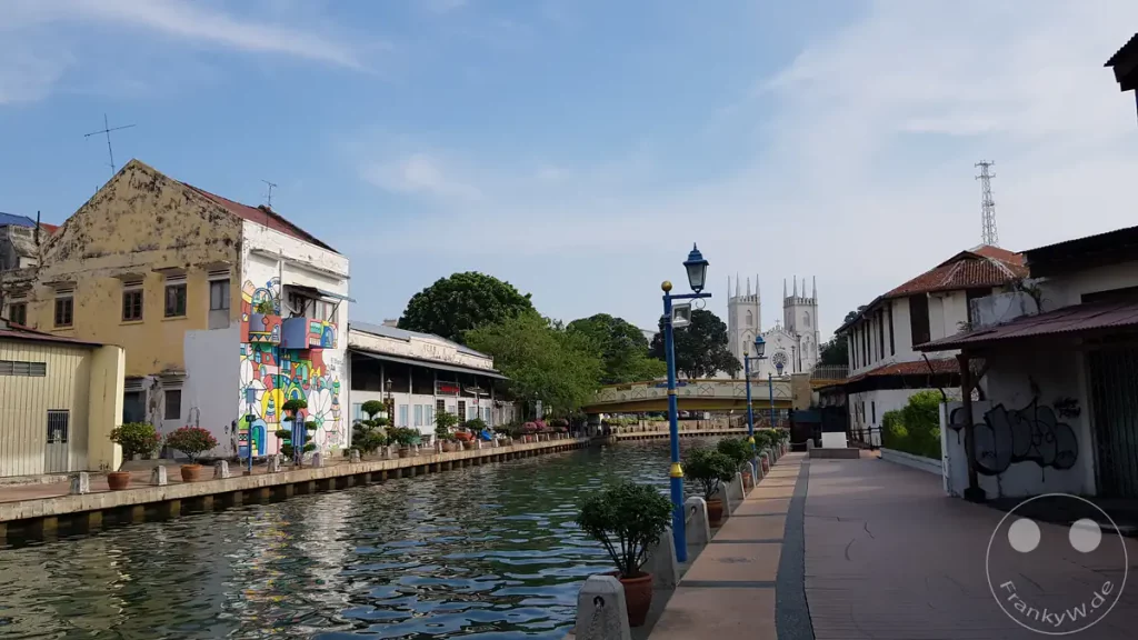 Blick entlang des Malakka-Flusses und der Uferpromenade in Malakka, Malaysia, mit farbenfrohen Wandmalereien an den Gebäuden, Straßenlaternen und den markanten weißen Türmen der St.-Francis-Xavier-Kirche im Hintergrund.