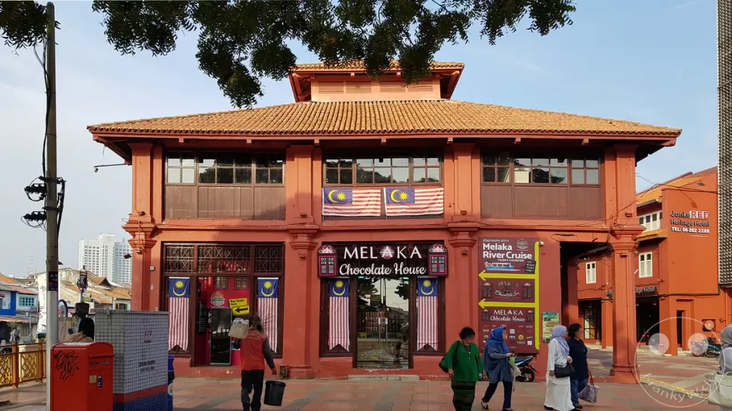 Fassade des historischen, rot gestrichenen Melaka Chocolate House am Roten Platz in Malakka, Malaysia, dekoriert mit malaysischen Flaggen und Passanten im Vordergrund.