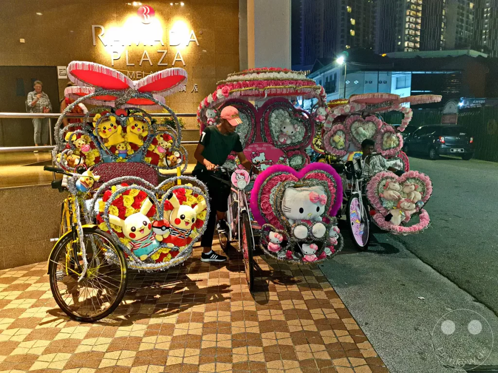 Nachtaufnahme vor dem Ramada Plaza Hotel in Malakka, Malaysia, mit aufwändig und farbenfroh mit Pikachu- und Hello-Kitty-Plüschfiguren, Lichterketten und künstlichen Blumen dekorierten Fahrrad-Rikschas.