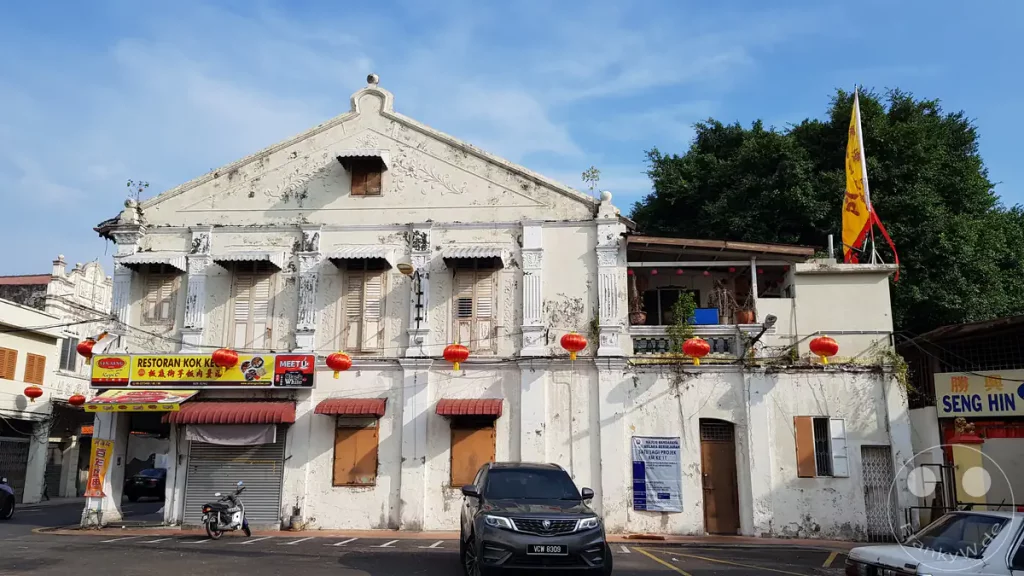 Fassade eines historischen, weiß getünchten Shophouses im Kolonialstil mit dem Restoran Kok Keong, traditionellen Fensterläden und roten Lampions an einer Straße in Malakka, Malaysia.