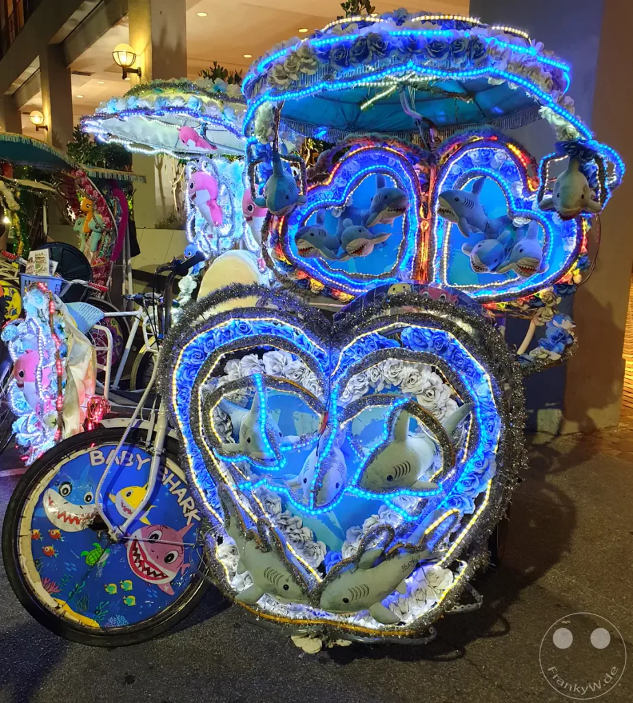 Nachtaufnahme einer traditionellen Fahrrad-Rikscha in Malakka, Malaysia, die aufwändig mit blau leuchtenden LED-Lichterketten, künstlichen weißen Rosen, Plüschhaien und Radkappen im „Baby Shark“-Design dekoriert ist.