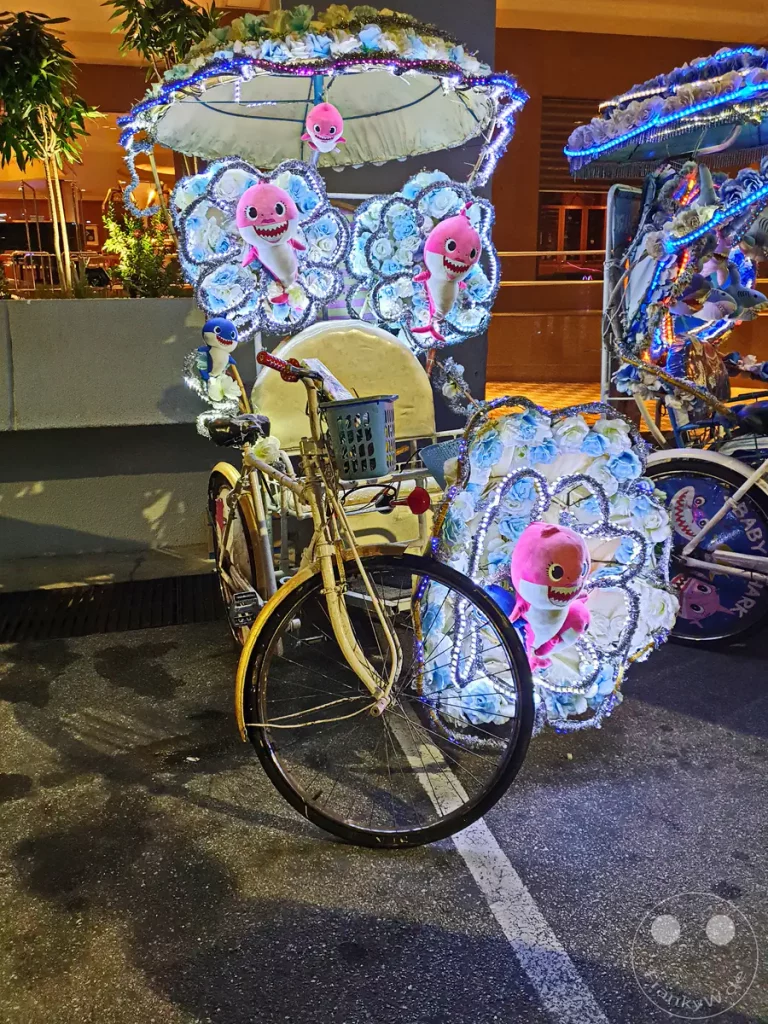 Nachtaufnahme einer traditionellen Fahrrad-Rikscha in Malakka, Malaysia, reich verziert mit rosa Plüschhaien im „Baby Shark“-Design, hellblau und weiß leuchtenden Lichterketten sowie künstlichen Rosenblüten.