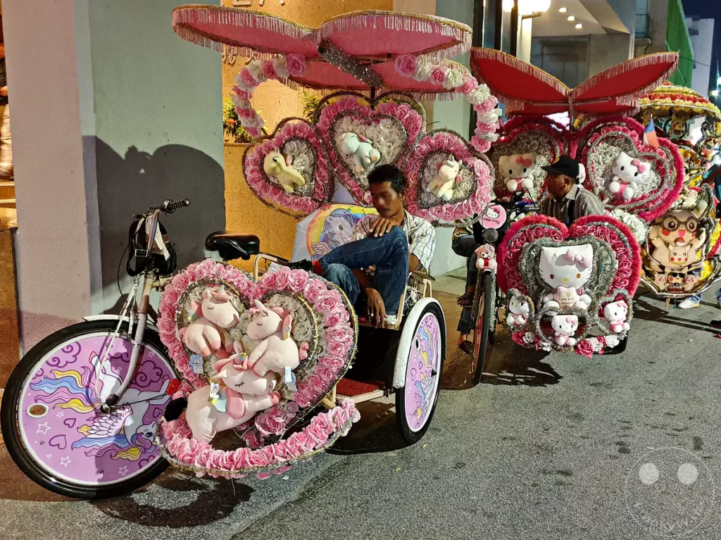 Nachtaufnahme von traditionellen Fahrrad-Rikschas in Malakka, Malaysia, aufwändig dekoriert mit rosafarbenen Kunstblumen, Plüsch-Einhörnern im „My Little Pony“-Stil und Hello-Kitty-Figuren, während die Fahrer am Straßenrand warten.