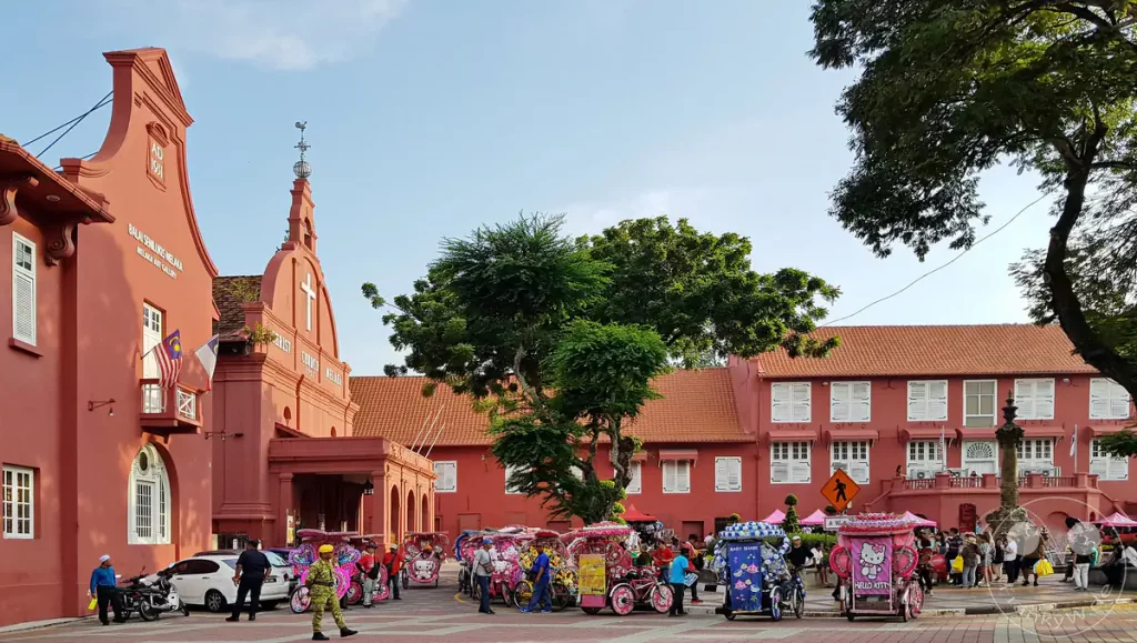 Blick auf den historischen Roten Platz in Malakka, Malaysia, mit den markanten roten Gebäuden der Christ Church und der Melaka Art Gallery, belebten Straßenszenen und farbenfroh dekorierten Fahrrad-Rikschas im Vordergrund.