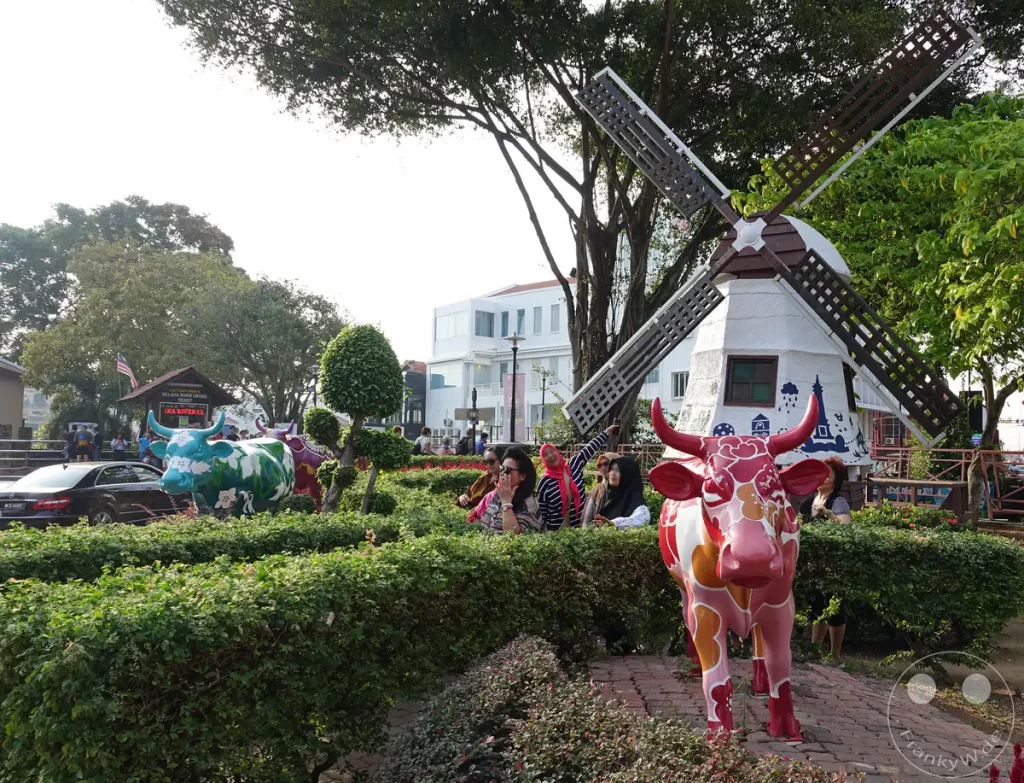 Blick auf eine kleine Parkanlage am historischen Dutch Square (Roten Platz) in Malakka, Malaysia, mit einer traditionellen holländischen Windmühle, farbenfroh verzierten Kuh-Skulpturen, grünen Hecken und Besuchern unter Bäumen.