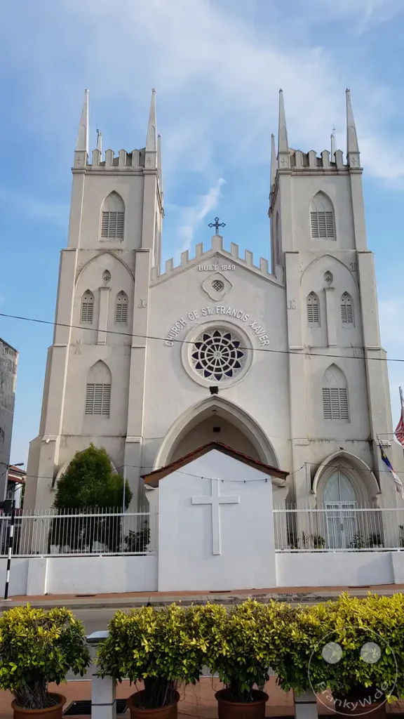 Vorderansicht der historischen, weißen St.-Francis-Xavier-Kirche in Malakka, Malaysia, mit ihren zwei neugotischen Türmen, einem großen Rosettenfenster und der Inschrift „Built 1849“ über dem Eingang.
