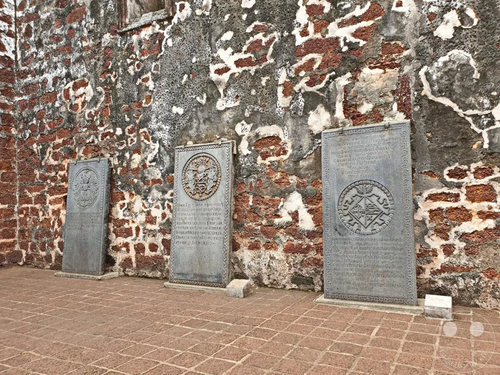 Drei historische, aufrecht stehende Grabplatten aus Stein mit eingemeißelten Wappen und Inschriften vor einer verwitterten, rotbraunen Laterit-Mauer in den Ruinen der St. Paul's Church in Malakka, Malaysia.
