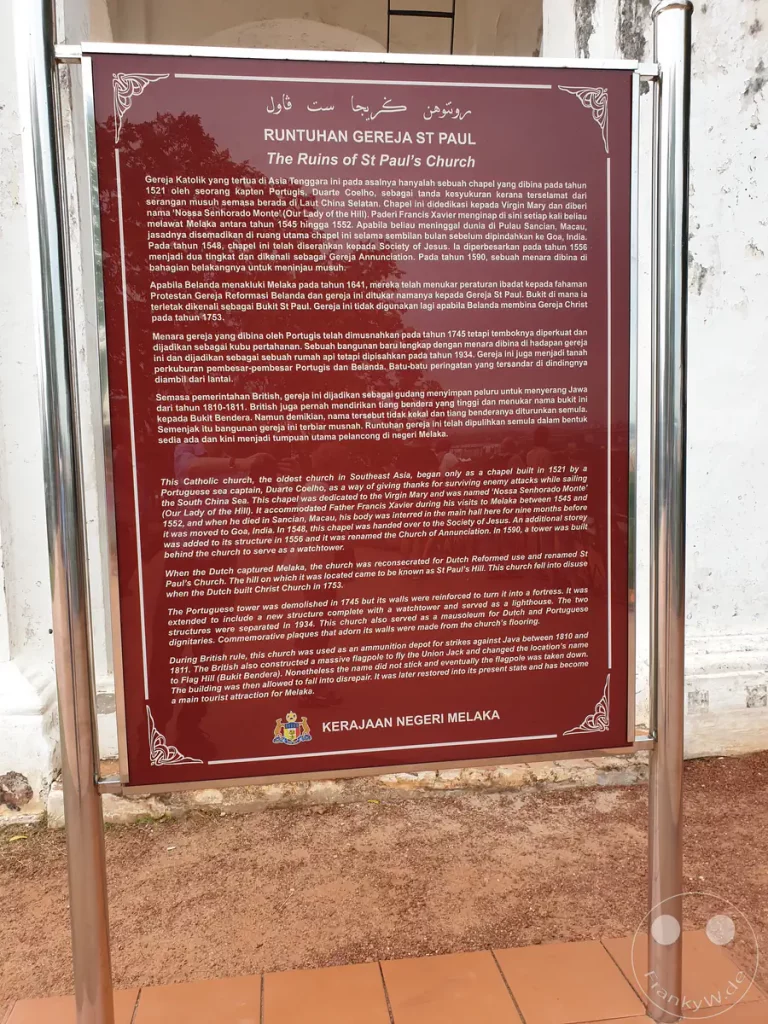 Eine rotbraune Informationstafel aus Metall an einer Steinmauer in Malakka, Malaysia, mit historischen Texten auf Malaiisch und Englisch über die Ruinen der St. Paul's Church, gekrönt vom Jawi-Schriftzug und dem Emblem der Regierung von Malakka.