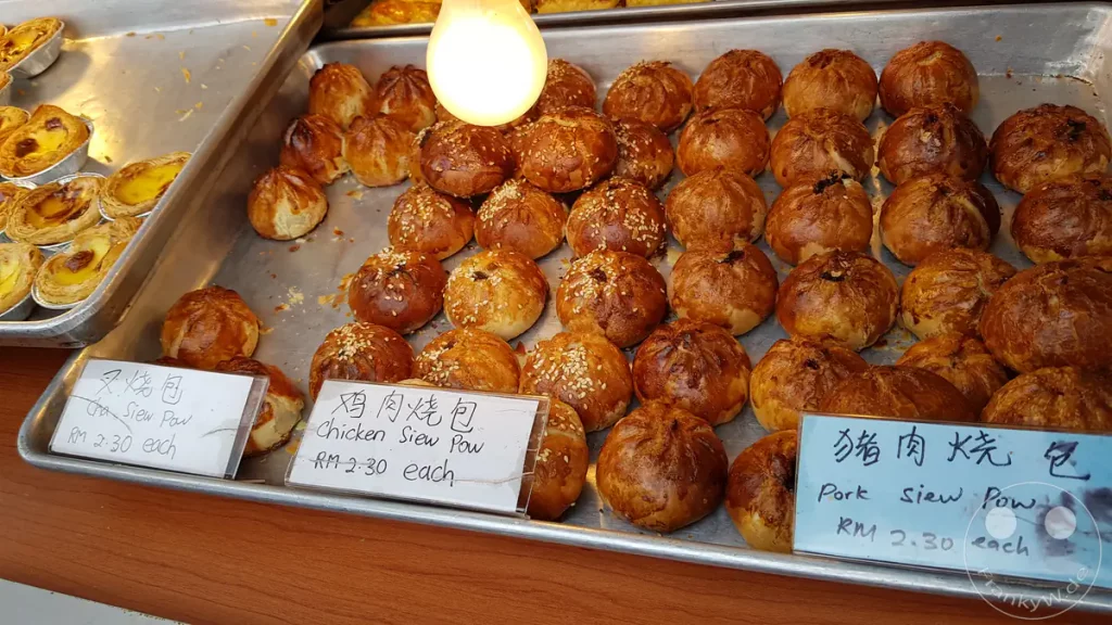 Frisch gebackene, goldbraune Siew Pow Teigtaschen mit Sesam auf einem Metallblech in einer malaysischen Auslage. Kleine Preisschilder beschriften die Sorten Huhn und Schweinefleisch.