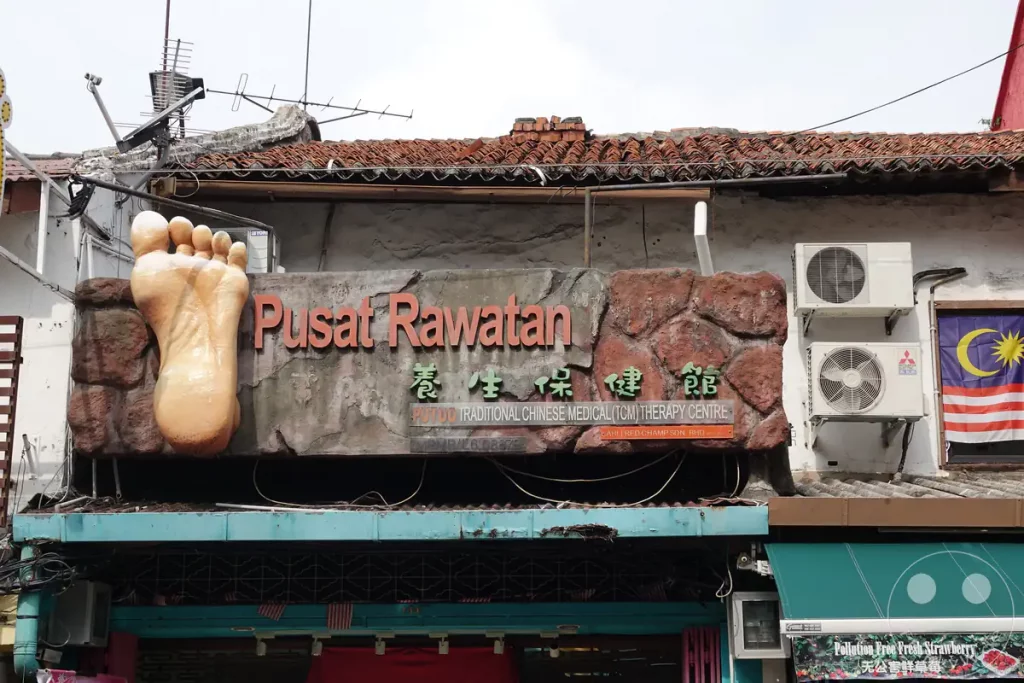 Ein auffälliges Werbeschild für ein Zentrum für Traditionelle Chinesische Medizin und Fußreflexzonenmassage (Pusat Rawatan) in der Stadt Melaka mit zwei großen plastischen Fuß-Symbolen über dem Eingang.