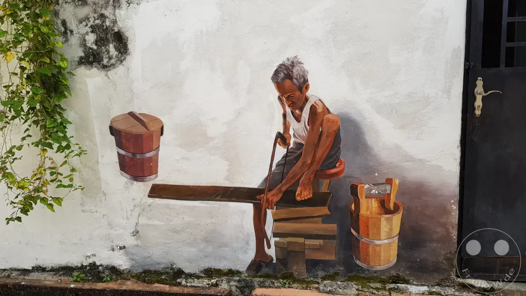 Ein realistisches Wandgemälde in der Stadt Melaka, das einen älteren Handwerker beim Sägen von Holz zeigt, umgeben von traditionellen Holzeimern an einer hellen Hauswand.