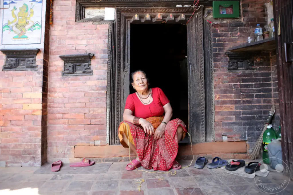 Eine ältere Frau sitzt auf den Stufen vor dem Eingang ihres traditionellen Hauses in Bhaktapur, Nepal. Sie trägt farbenfrohe Kleidung und ist von den Schuhen umgeben, die vor dem Eingang stehen.
