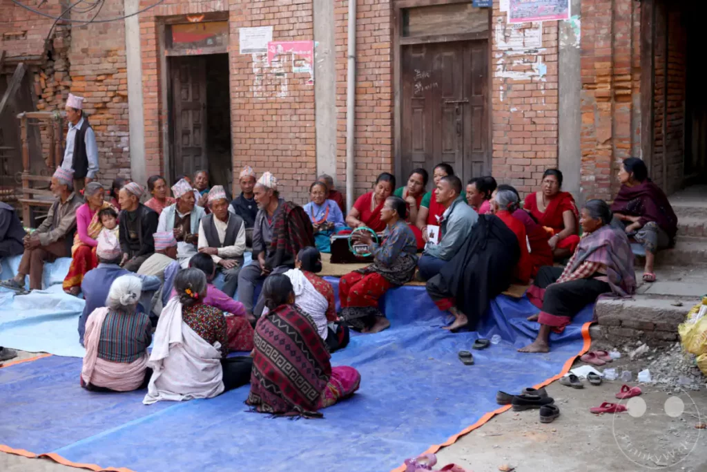 Eine Gruppe von älteren Männern und Frauen sitzt auf einer blauen Plane vor einem Backsteinhaus in Bhaktapur, Nepal. Sie tragen traditionelle Kleidung und einige von ihnen Kopfbedeckungen. Die Szene wirkt ruhig und gemeinschaftlich.