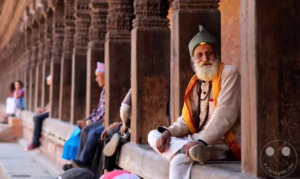 Ein älterer Mann mit Bart und traditioneller Kleidung sitzt im Freien vor kunstvoll geschnitzten Holzsäulen in Bhaktapur, Nepal. Die ruhige und nachdenkliche Pose des Mannes in der historischen Umgebung erzeugt eine gelassene Atmosphäre.