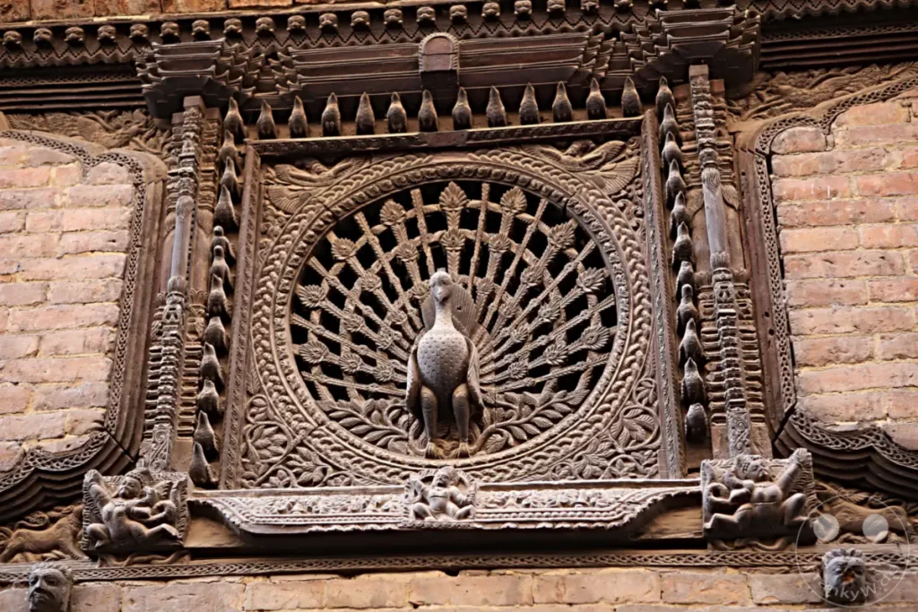 Ein aufwendig geschnitztes hölzernes Fenster im Stil eines Pfauenschwanzes befindet sich in Bhaktapur, Nepal. Die detaillierte Handwerkskunst und die reiche Tradition sind auf dem Bild deutlich erkennbar. Das Fenster ist mit zahlreichen kleinen Figuren und Ornamenten verziert.