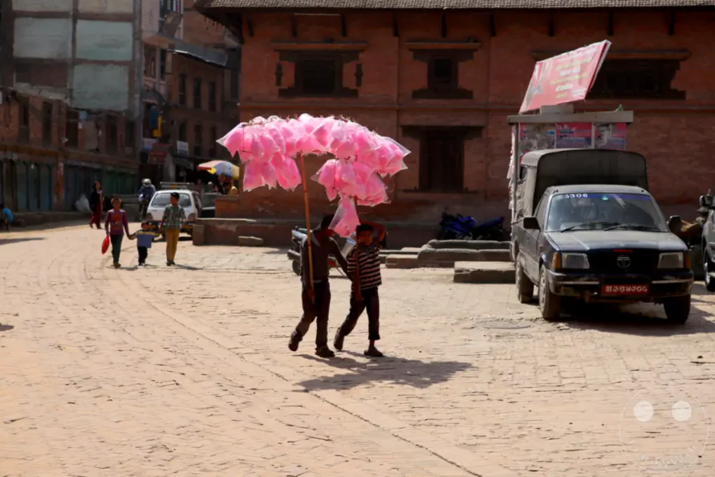 Zwei junge Jungen tragen in Bhaktapur, Nepal, eine improvisierte Stange mit mehreren bunten Zuckerwatte-Portionen. Die Szene spielt sich auf einem gepflasterten Platz ab, mit traditionellen Backsteingebäuden im Hintergrund und einer belebten Marktatmosphäre.