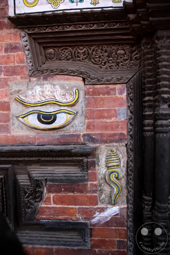 Ein detaillierter Blick auf die kunstvollen Holzschnitzereien und ein auffälliges Augenmotiv, das an eine Ziegelwand eines Tempels in Bhaktapur angebracht ist. Die komplexe Handwerkskunst und die traditionellen Symbole erzählen von der reichen Kultur und Spiritualität der Region.