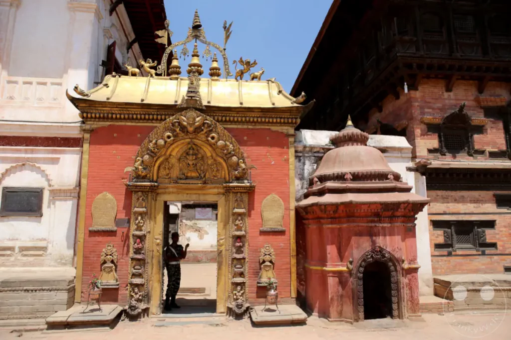 Eine goldverzierte Toranlage mit einer Wache in militärischer Uniform, die davor steht. Nebenan befindet sich ein kleiner roter Schrein. Die Szene spielt sich auf einem sonnigen Platz in Bhaktapur, Nepal, ab.