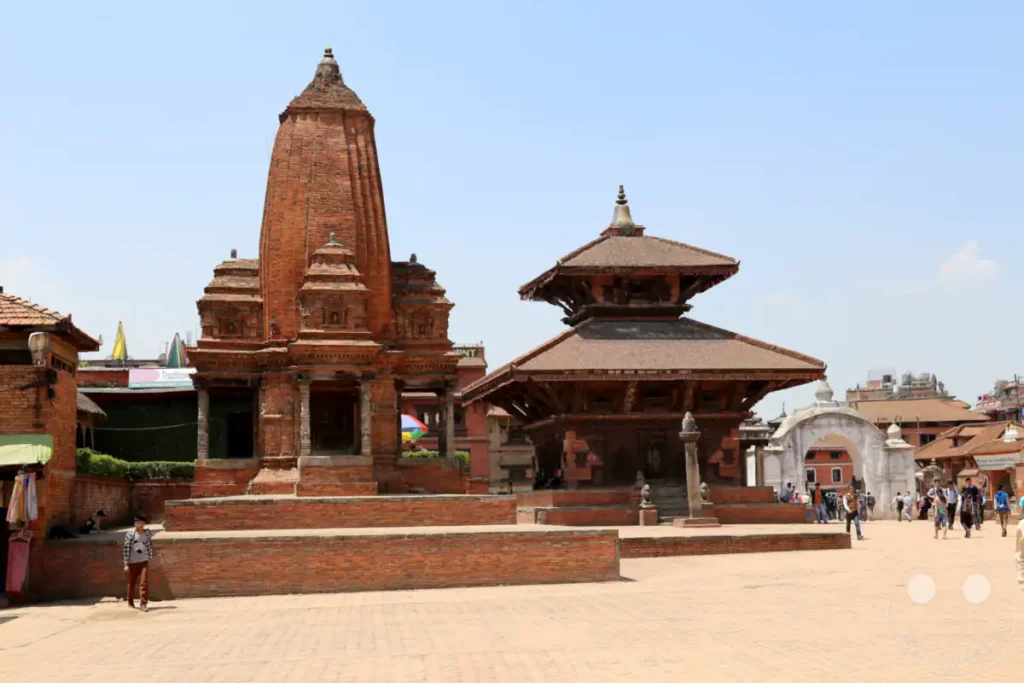 Eine Aufnahme des Tempelkomplexes Nyatapola in Bhaktapur, Nepal. Touristen schlendern über den gepflasterten Platz, während im Hintergrund die charakteristische Pagodenarchitektur zu sehen ist. Das Bild vermittelt eine friedliche und kulturelle Atmosphäre.