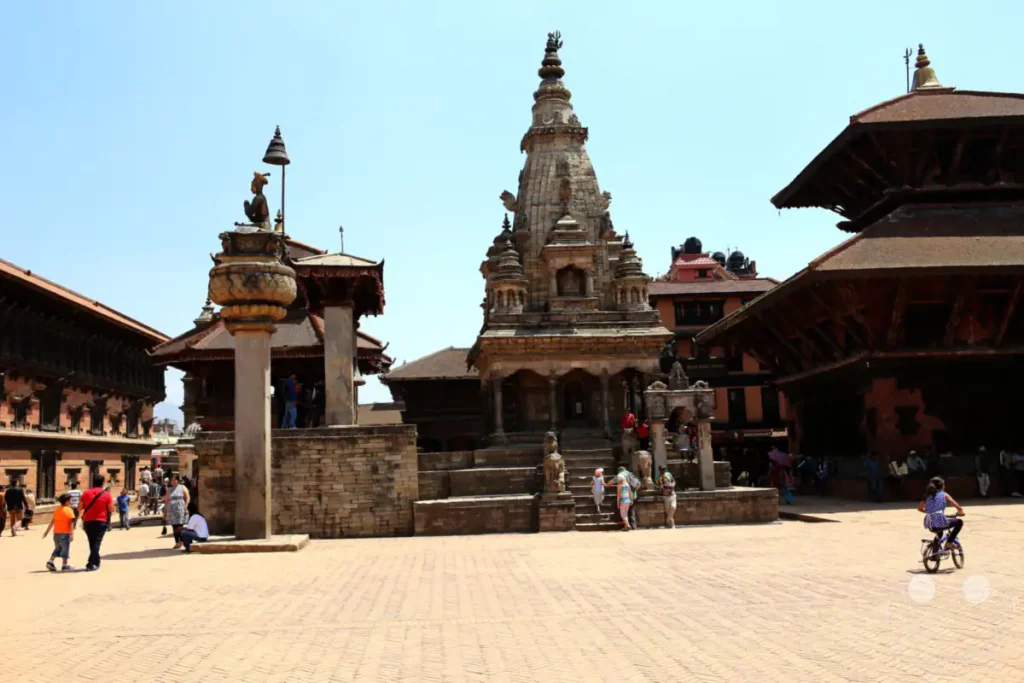 Der Tempelplatz von Bhaktapur zeigt traditionelle nepalesische Architektur und Steinmetzarbeiten. Menschen bewegen sich durch den belebten Platz, was auf eine aktive kulturelle Stätte hinweist. Die Szene vermittelt einen Eindruck von lokaler Lebensweise und historischem Erbe.